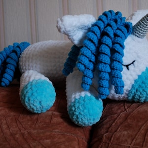 May include: A white crocheted unicorn soft toy with blue yarn for the mane and tail. The unicorn is lying down with its eyes closed.