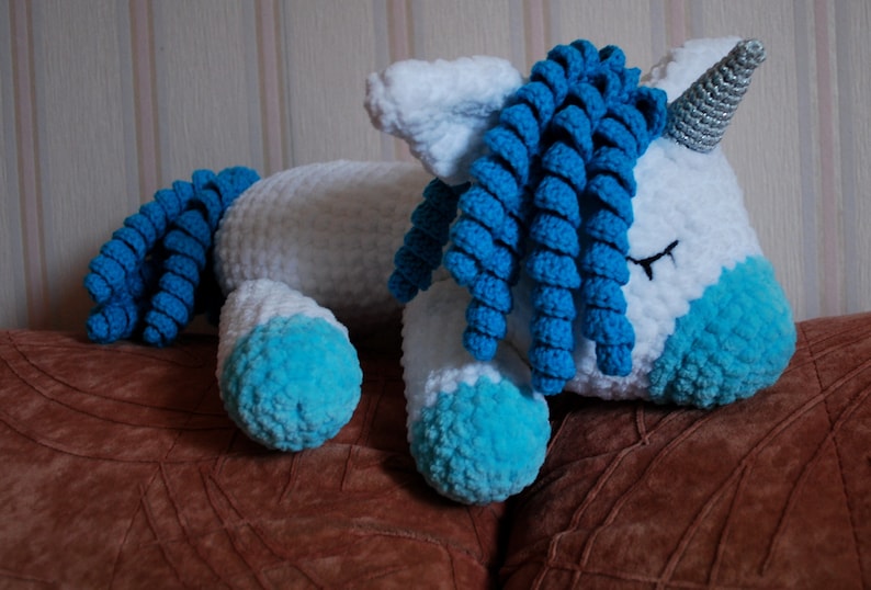 May include: A white crocheted unicorn soft toy with blue yarn for the mane and tail. The unicorn is lying down with its eyes closed.