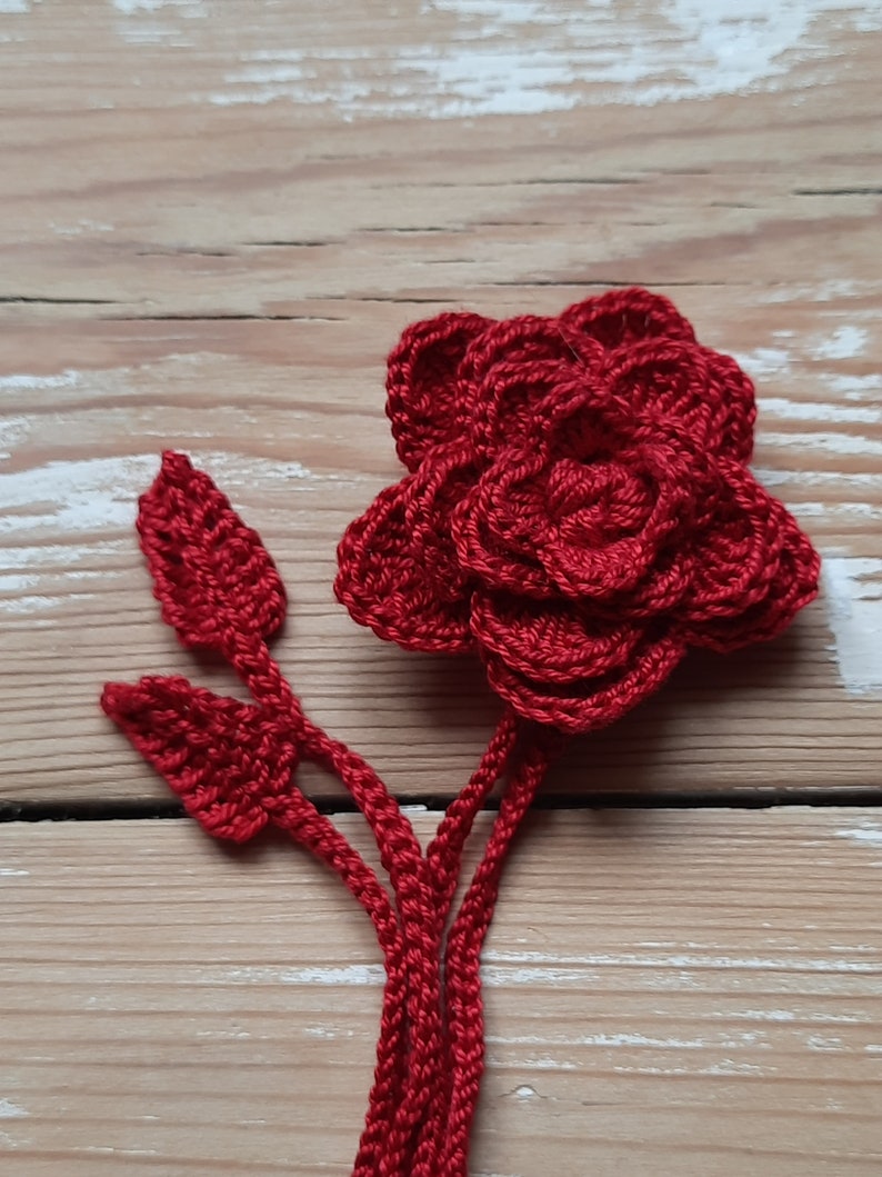 May include: A crocheted red rose with a stem and leaves. The rose is made of yarn and has a layered, textured appearance.