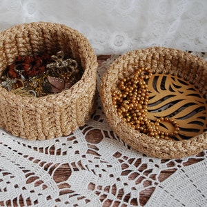 Pu&ograve; includere: Due cesti intrecciati, uno pieno di gioielli e l'altro con una collana d'oro e una foglia di legno decorativa. I cesti sono su un pizzo bianco all'uncinetto.
