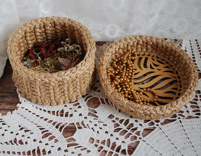 Pu&ograve; includere: Due cesti intrecciati, uno pieno di gioielli e l'altro con una collana d'oro e una foglia di legno decorativa. I cesti sono su un pizzo bianco all'uncinetto.