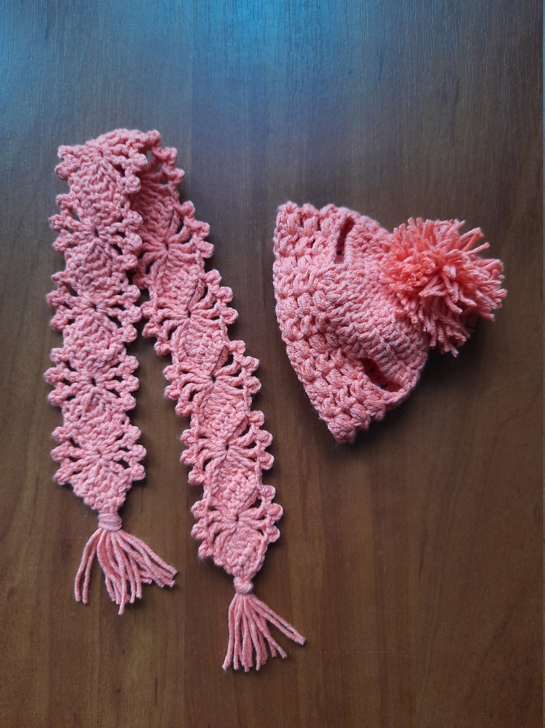 May include: A pink crocheted hat with a pom-pom and a matching scarf with tassels.