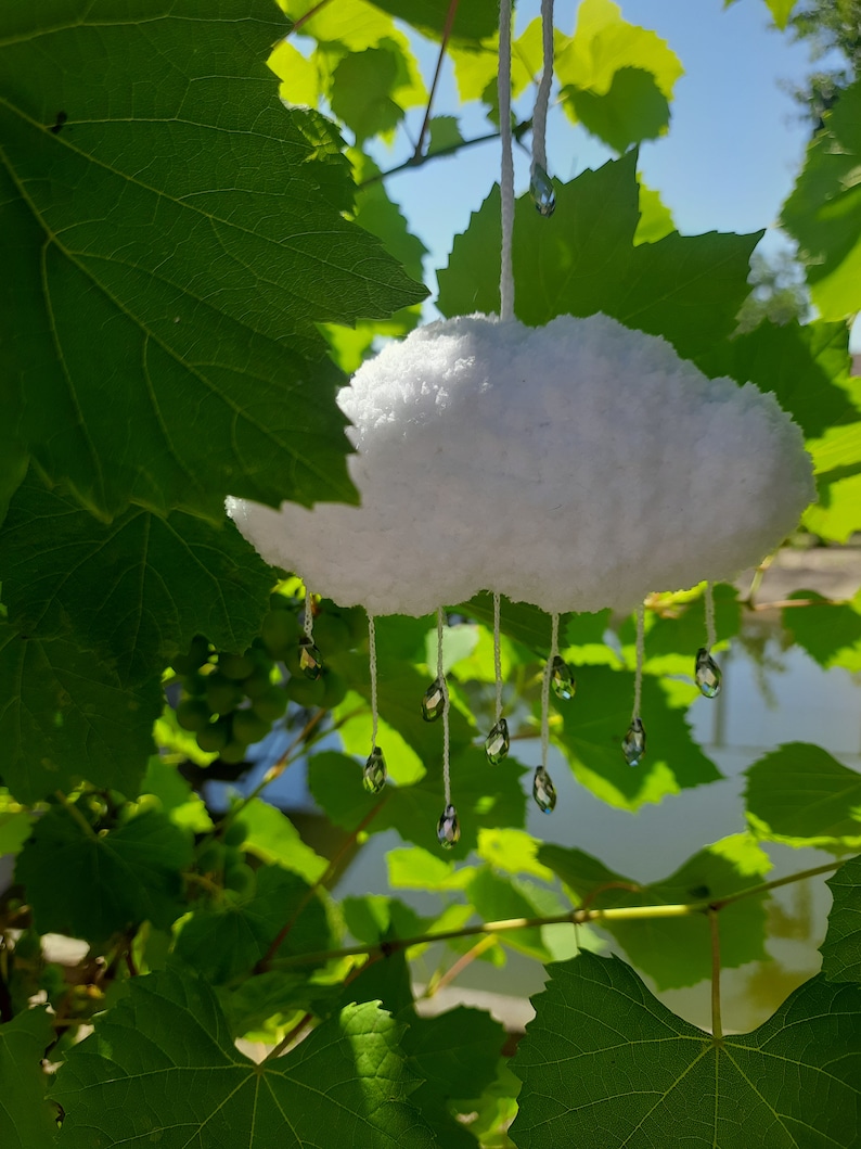 Puede incluir: Decoraci&oacute;n de nube blanca y esponjosa con gotas de cristal colgantes. La nube est&aacute; suspendida por un hilo blanco, sobre un fondo de hojas verdes y luz solar. Ideal para la decoraci&oacute;n del hogar.