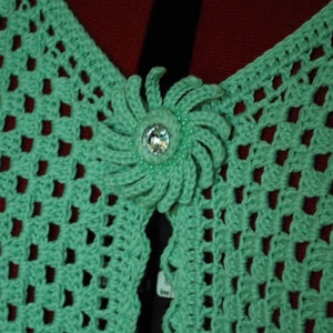 May include: A green crocheted shawl with a decorative flower-shaped button in the centre.