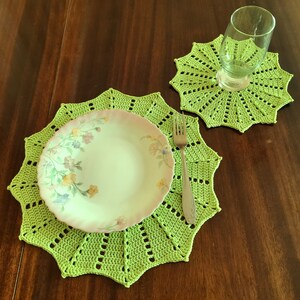 May include: A green crocheted placemat with a floral patterned plate and a silver fork on top. A smaller green crocheted coaster is in the background.