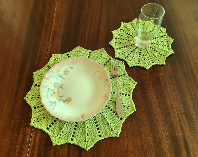 May include: A green crocheted placemat with a floral patterned plate and a silver fork on top. A smaller green crocheted coaster is in the background.