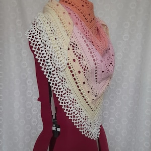 May include: A handmade crochet shawl in a gradient of colors, from white to peach. The shawl is draped over a mannequin, showcasing its intricate openwork design. The edges are finished with a delicate, lacy trim, adding a touch of elegance to the accessory.