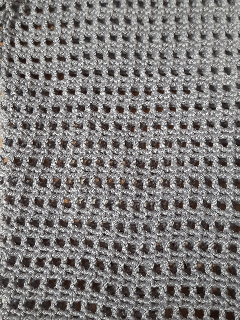 K&ouml;nnte beinhalten: Nahaufnahme eines grau geh&auml;kelten Stoffes mit einem netzartigen Muster. Das Garn ist einfarbig mittelgrau, und die Maschen bilden eine Reihe gleichm&auml;&szlig;ig verteilter quadratischer &Ouml;ffnungen. Die Textur ist sichtbar, was auf einen handgefertigten Artikel hindeutet.