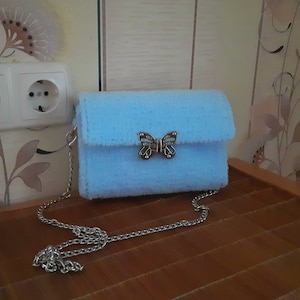 Puede incluir: Un bolso de peluche azul claro con un cierre de mariposa plateado y una correa de cadena plateada.
