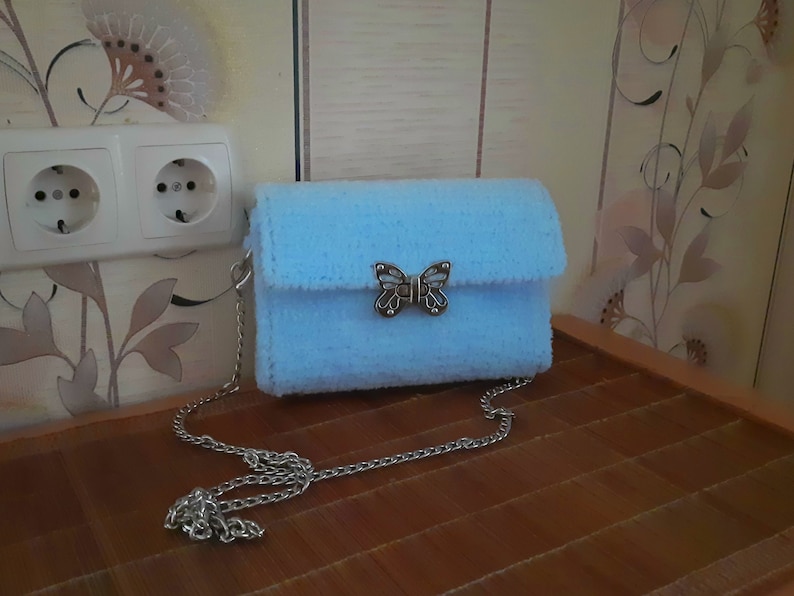 Puede incluir: Un bolso de peluche azul claro con un cierre de mariposa plateado y una correa de cadena plateada.