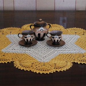 May include: A brown ceramic tea set with a teapot and two cups and saucers, set on a yellow and white crocheted doily.