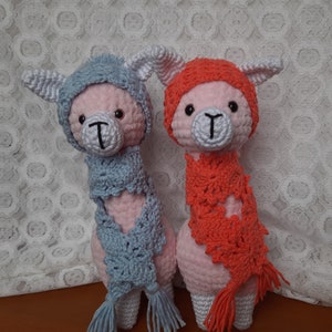 May include: Two crocheted llama toys wearing hats and scarves. The llama on the left is wearing a light blue hat and scarf, while the llama on the right is wearing an orange hat and scarf.