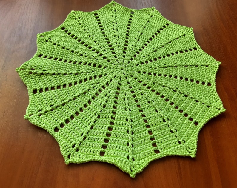 May include: A green crocheted doily with a starburst pattern. The doily is made with a single crochet stitch and has a scalloped edge.