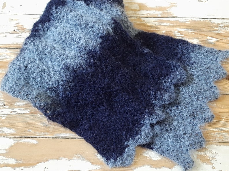 May include: A soft, blue and gray knitted scarf with a fuzzy texture. The scarf is made of a gradient yarn that transitions from a light blue to a dark blue.