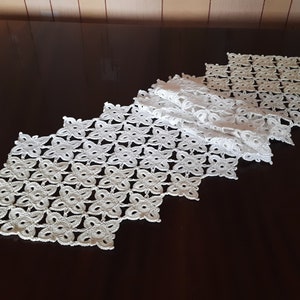 May include: White crocheted table runner with a diamond pattern. The runner is made of delicate, openwork stitches and has a scalloped edge.