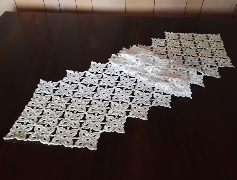 May include: White crocheted table runner with a diamond pattern. The runner is made of delicate, openwork stitches and has a scalloped edge.