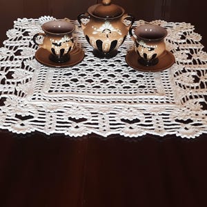 May include: A white crocheted doily with a delicate floral pattern sits on a dark wooden table. Three small brown ceramic cups with floral designs and a matching brown ceramic teapot are arranged on the doily.
