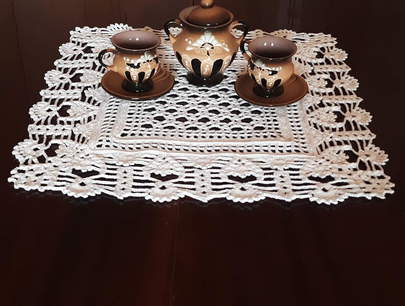 May include: A white crocheted doily with a delicate floral pattern sits on a dark wooden table. Three small brown ceramic cups with floral designs and a matching brown ceramic teapot are arranged on the doily.