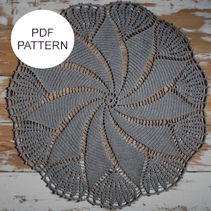 Puede incluir: Un tapete gris tejido a ganchillo con un diseño en espiral y un borde festoneado. El tapete es circular y presenta un patrón detallado. El texto "PDF PATTERN" está en un círculo blanco en la esquina superior izquierda. El tapete está sobre una superficie de madera envejecida.