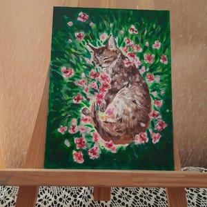 Puede incluir: Una pintura de un gato durmiendo en un campo de flores rosas. El gato es marr&oacute;n y blanco y tiene los ojos cerrados. Las flores est&aacute;n rodeadas de hierba verde.