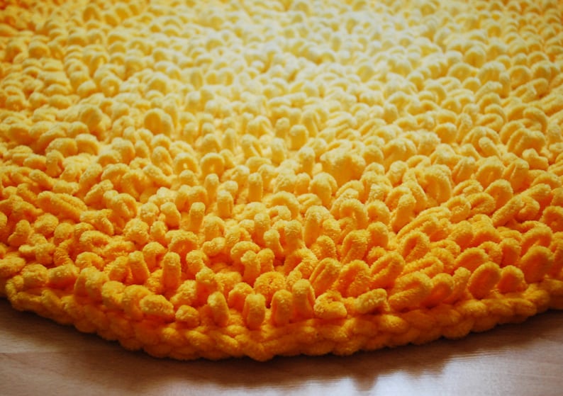 May include: A close-up of a yellow bath mat with a gradient pattern. The mat is made of soft, fluffy material and has a round shape.