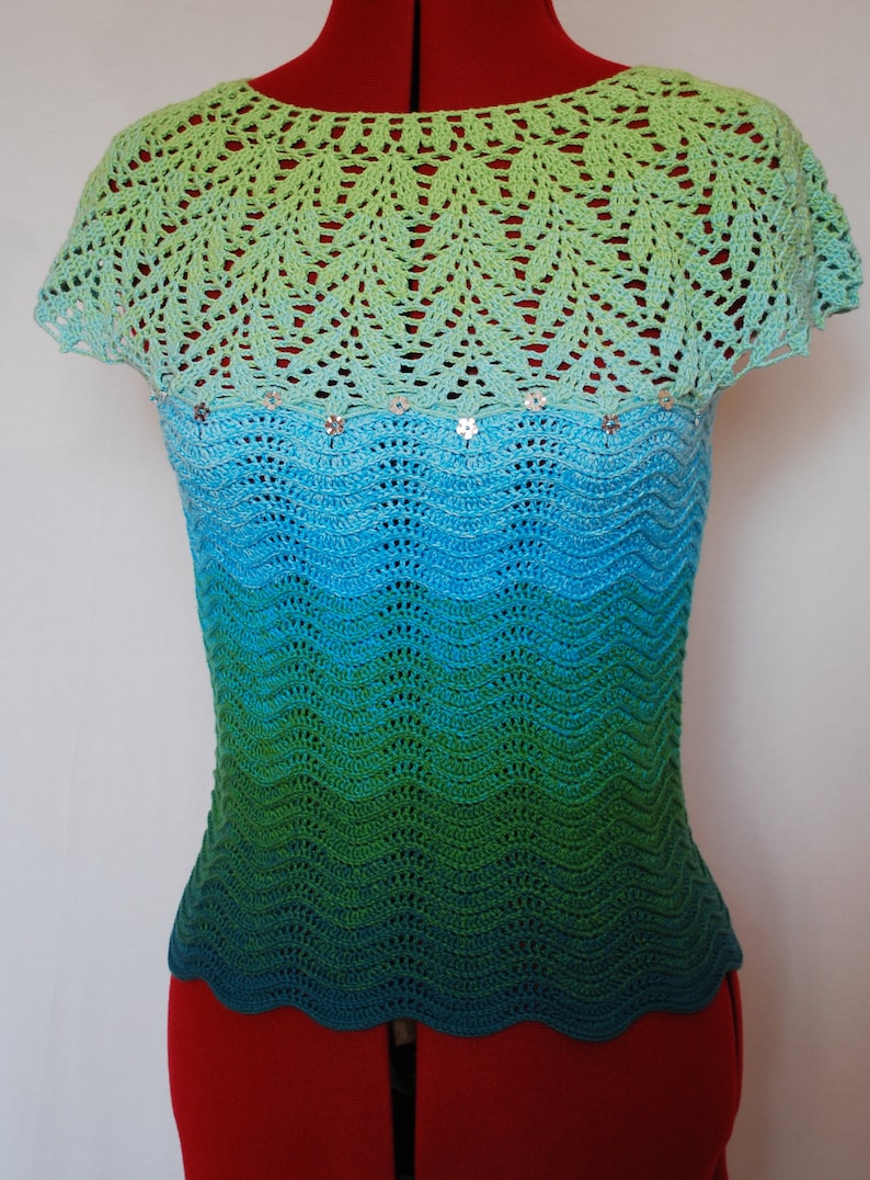 May include: A crocheted top with a green, blue, and turquoise ombre design. The top has a scalloped hem and a delicate crocheted lace neckline. The top is adorned with small, silver sequins.