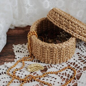 Pu&ograve; includere: Una scatola in crochet con coperchio, realizzata in filato beige naturale. La scatola &egrave; piena di gioielli e perline dorate. La scatola &egrave; appoggiata su un pizzo bianco all'uncinetto.