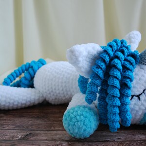 May include: A white crocheted unicorn with a blue mane and tail. The unicorn is lying down with its eyes closed. The unicorn has a silver horn.