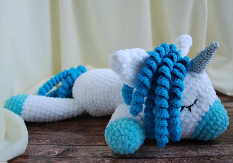 May include: A white crocheted unicorn with a blue mane and tail. The unicorn is lying down with its eyes closed. The unicorn has a silver horn.