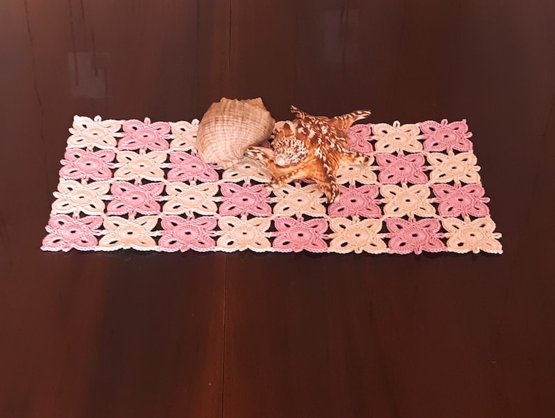 May include: A crocheted pink and beige table runner with a seashell on top.