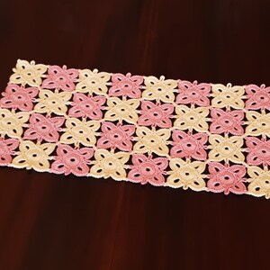 May include: A crocheted table runner with a pink and cream floral pattern. The runner is made of delicate yarn and has a square shape.
