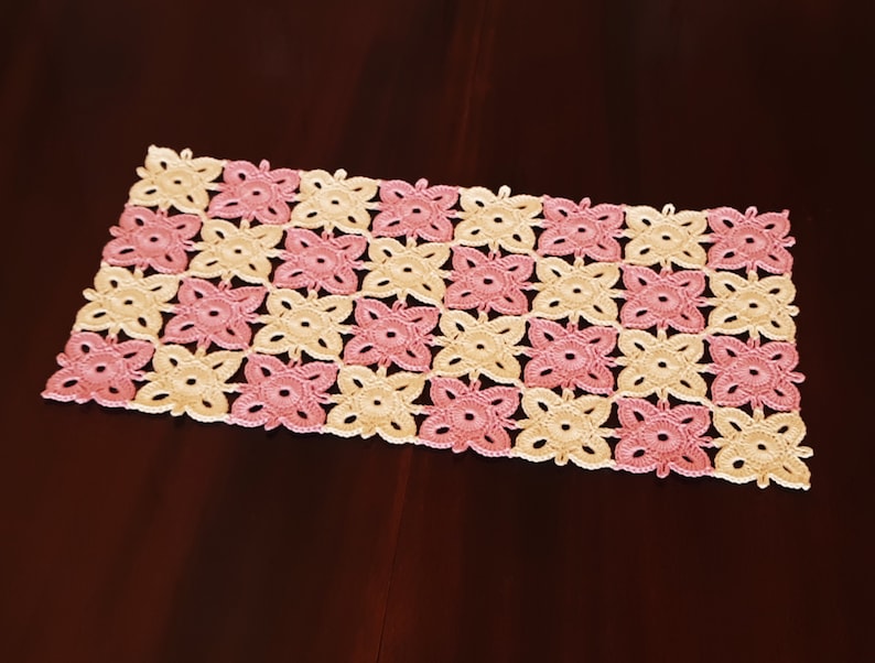 May include: A crocheted table runner with a pink and cream floral pattern. The runner is made of delicate yarn and has a square shape.