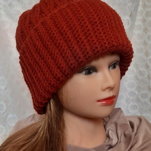 May include: A red knitted beanie hat with a ribbed texture. The hat is worn on a mannequin with long brown hair.