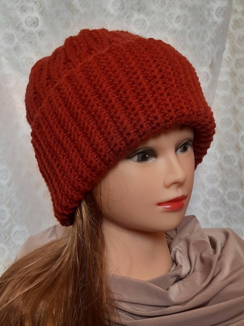May include: A red knitted beanie hat with a ribbed texture. The hat is worn on a mannequin with long brown hair.