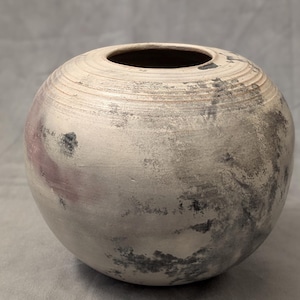 May include: A white ceramic vase with a round shape and a textured surface. The vase has a subtle grey and pink colour variation.