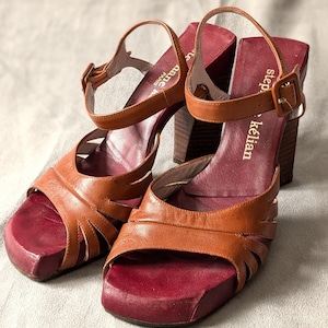 May include: A pair of brown leather heeled sandals with a burgundy platform. The sandals have a strap across the ankle and a buckle closure. The brand name "step & kelian" is printed on the strap.