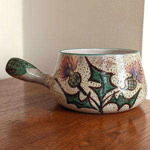 May include: A white ceramic serving dish with a green, brown, and pink floral design. The dish has a handle and is ideal for serving sauces or dips.