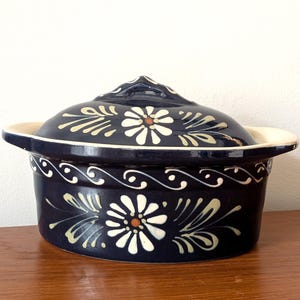 Può includere: Una casseruola in ceramica blu con coperchio, con un motivo floreale bianco con accenti gialli. La casseruola ha una forma arrotondata e due manici.