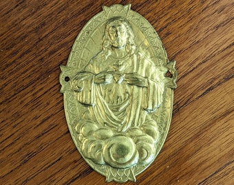 May include: A gold-colored metal religious medallion depicting Jesus Christ with a heart in his hand. The medallion is oval-shaped and has a decorative border.