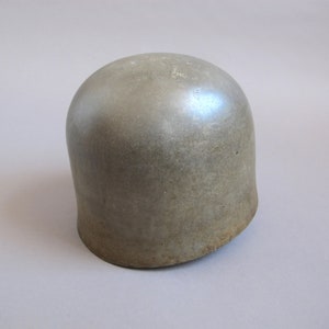 Könnte beinhalten: Ein grauer Metallhelm mit abgerundeter Oberseite und leicht abgewinkeltem Rand. Der Helm hat ein abgenutztes, verwittertes Aussehen.