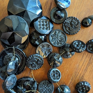 May include: A collection of black glass buttons with various designs and shapes. Some buttons have a shiny finish, while others have a matte finish.