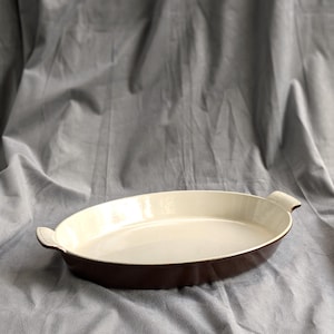 May include: An oval-shaped baking dish with a white interior and two handles. The dish is made of enamelled cast iron and is brown in colour.