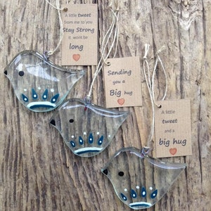 May include: Three clear glass bird ornaments with painted details and attached tags. The tags have messages like "Stay Strong" and "Big hug". The ornaments are hanging on a wooden surface.