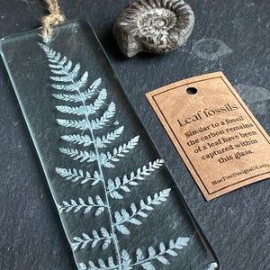 Puede incluir: Un marcapáginas rectangular de vidrio transparente con un diseño de helecho y una cuerda de yute. Junto a él, un pequeño fósil gris oscuro y una etiqueta marrón con el texto "Leaf fossils". La etiqueta explica que los restos de carbono de una hoja están capturados en el vidrio.