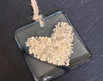 May include: A clear, square pendant with a heart-shaped design filled with gold-colored flakes. The pendant is attached to a natural twine cord. The background is a dark, textured surface.