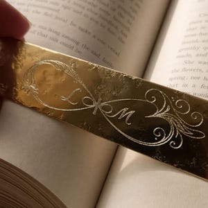 Può includere: Un segnalibro rettangolare in metallo color oro con un simbolo dell'infinito inciso. Il centro dell'anello dell'infinito contiene le lettere corsive L e M, circondate da intricati motivi a filigrana sulla superficie lucida.