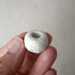 Puede incluir: Un pequeño objeto en forma de rosquilla con una superficie rugosa y texturizada. El objeto es de color gris claro y tiene un agujero en el centro. Lo sostiene la mano de una persona.