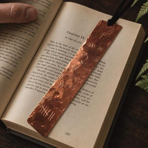Op de afbeelding: Een rechthoekige koperen boekenlegger met een gehamerd, getextureerd oppervlak. De boekenlegger is voorzien van een gegraveerd kruissymbool bij de bovenkant en een reeks verticale streepjes bij de onderkant. Een zwart koord is bevestigd door een gat aan de bovenrand.
