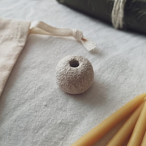 Puede incluir: Una pequeña cuenta redonda de piedra beige con un agujero central, sobre una superficie de lino blanco. La cuenta tiene una apariencia texturizada y porosa. Otros elementos incluyen una bolsa beige con cordón, un artículo verde enrollado y varias velas amarillas.