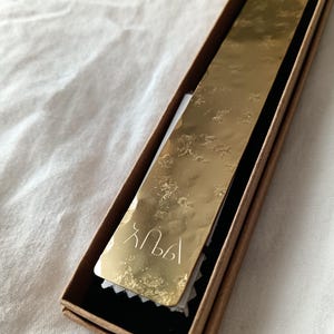 Peut inclure: Un marque-page rectangulaire en m&eacute;tal dor&eacute; avec une texture de surface martel&eacute;e et r&eacute;fl&eacute;chissante. Le marque-page pr&eacute;sente des caract&egrave;res grav&eacute;s en bas et est attach&eacute; &agrave; un fin cordon vert, pr&eacute;sent&eacute; dans une bo&icirc;te cadeau rectangulaire en carton brun.